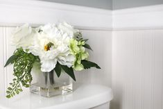 white flowers are in a square vase on the toilet