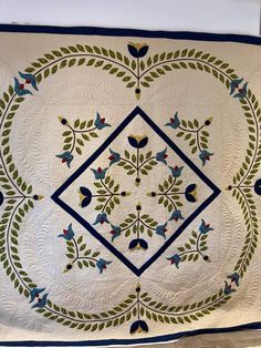 a white quilt with blue and green designs on the front, hanging from a wall