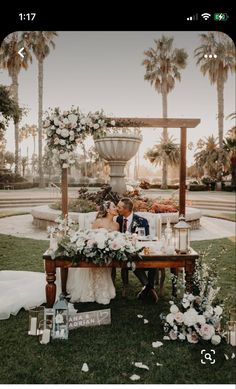 a bride and groom sitting at a table in front of a fountain surrounded by flowers