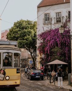 a yellow trolley car driving down a street next to tall buildings with purple flowers on them