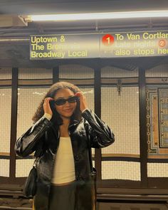 a woman wearing sunglasses standing in front of a subway station sign with her hand on her head