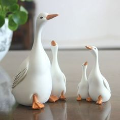 three white ducks standing next to each other on top of a wooden table in front of a potted plant