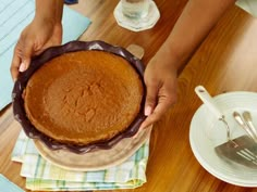 a person holding a pie on top of a wooden table next to plates and utensils