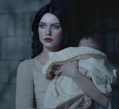 a woman with blue eyes holding a baby in a white dress and gold chain around her neck