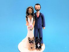 a wedding cake topper with a bride and groom holding two small dogs, on a blue background