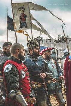 men dressed in medieval clothing standing next to each other with flags flying behind them and looking at the ground