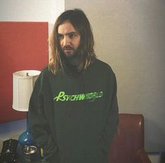 a man with long hair wearing a black hoodie standing in front of a red couch