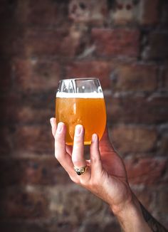 a person holding up a glass of beer in front of a brick wall with a ring on it's finger