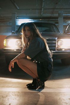 a woman squatting down on her skateboard in a parking garage with a car behind her