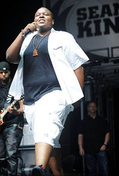 a man in white shorts and black shirt singing on stage with another man standing behind him