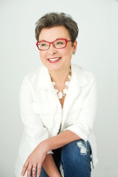 a woman with glasses is posing for a photo in front of a white background and has her hands on her knees