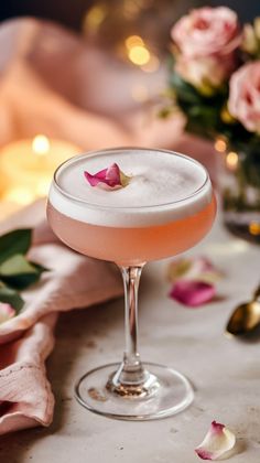 a pink cocktail with rose petals in a glass on a table next to candles and flowers