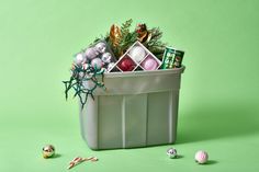 a trash can filled with assorted christmas decorations on a green background next to balls and tinsel