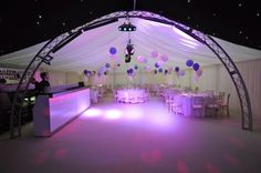 a party tent set up with white tables and purple balloons hanging from the ceiling above it