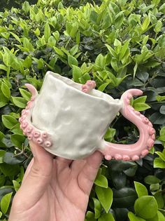 a hand holding a ceramic cup with an octopus decoration on the outside, and green bushes in the background