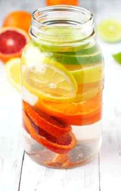 a glass jar filled with sliced fruit on top of a white wooden table next to oranges and limes