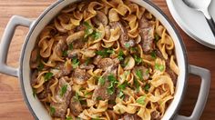 a pot filled with pasta and meat on top of a wooden table next to silverware