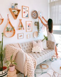 a woman hanging pictures on the wall above a couch in a room with plants and other decorations