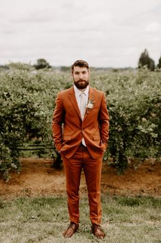 a man in an orange suit and tie standing in front of some bushes with his hands in his pockets