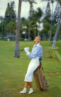 a woman in white dress holding a golf bag on grass with palm trees behind her