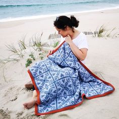 a woman sitting on the beach wrapped in a blue and white blanket with an orange border