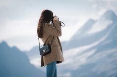 a woman standing on top of a mountain holding a camera up to her face and looking into the distance