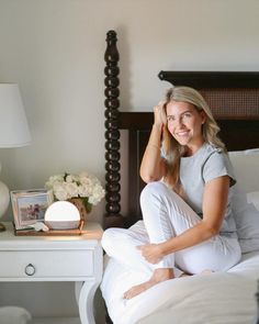 a woman sitting on top of a bed next to a night stand and table with flowers