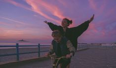 two people riding bikes on the beach at sunset with pink clouds in the sky above them