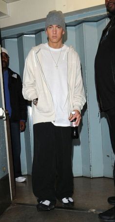 a young man in white jacket and black pants standing next to a wall with other people