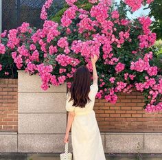 a woman in a white dress is holding her hand up to the pink flower bush