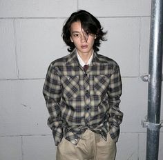 a young man standing in front of a wall wearing a plaid shirt and khaki pants