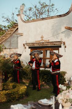 a group of men in mexican attire playing instruments on the grass near a building with flowers