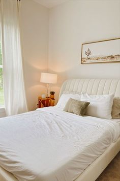 a white bed sitting under a window next to a lamp on a table in front of a window