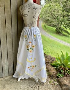 "white cotton (? linen ) flared skirt with appliqué yellow flowers & embroidery bias cut and with cotton crochet at the hemline unlined SIZE - the elastic waist stretches from 66 cm /26\" to 99 cm / 39\" ~ fastens at the back ~ length averages 107 cm / 42\" mannequin is size small / m gentle wash" White Embroidered Maxi Skirt For Spring, Fitted White Skirt With Embroidered Hem, Yellow Bottoms With Floral Embroidery For Summer, Yellow Floral Embroidery Bottoms For Summer, Yellow Floral Embroidery Bottoms For Spring, Spring Yellow Bottoms With Floral Embroidery, Yellow Embroidered Summer Bottoms, Yellow Embroidered Bottoms For Summer, White Long Skirt For Garden Party