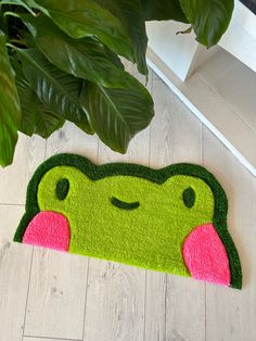 a green frog door mat with pink lips on the floor next to a potted plant