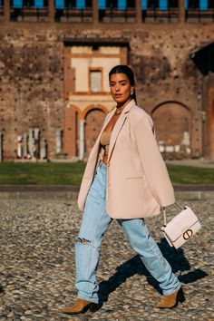 a woman walking across a cobblestone street holding a white purse and wearing jeans