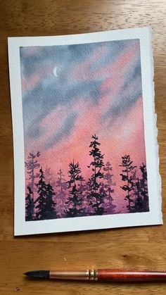 a watercolor painting of trees with the moon in the sky above them and a paintbrush next to it on a wooden table