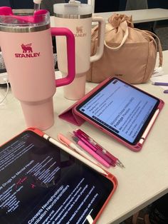 the tablet is next to a pink cup and coffee mug on the table with other items