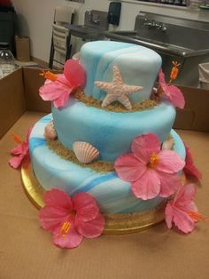 a three tiered blue cake with pink flowers on the bottom and starfish on top