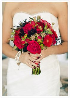 a woman in a wedding dress holding a bouquet of red roses and greenery on her hands