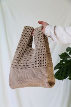 a hand is holding a crocheted bag in front of a potted plant