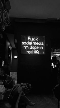 black and white photograph of people sitting at tables in front of a sign that reads f k social media, i'm dope in real life