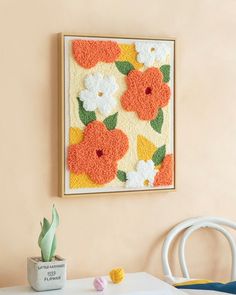 an orange and white flowered wall hanging on the wall next to a potted plant