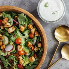 a wooden bowl filled with salad next to two spoons and a jar of ranch dressing