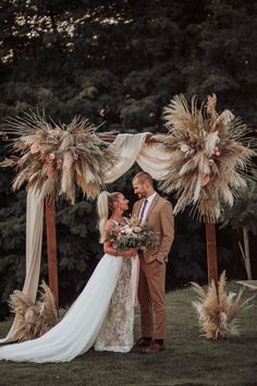 a bride and groom are standing under an arch decorated with pamodia flowers, grass and feathers