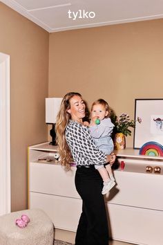 a woman holding a baby in her arms and standing next to a dresser with toys on it