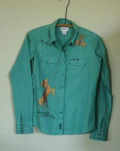 wrangler cowgirl shirt. size S but it fit's more like an XS. teal cotton with a gold metallic thread. snap closure down the front and at the cuffs. gold transfers of a horse and guitar's. star and circle rivet embellishments. 'cowgirls rock' embroidered on the lower right side. western cut at the yoke front and back. very good vintage condition, no stains, holes or flaws. by wrangler. shoulder width  15bust at armpit 36sleeve  24length  23.25 Western Style Shirt For Rodeo In Spring, Western Style Relaxed Fit Button-up Tops, Trendy Fitted Tops For Rodeo, Western Style Cotton Shirt For Spring, Fall Cotton Fitted Shirt, Fitted Cotton Shirt For Fall, Country Style Button-up Cotton Tops, Cotton Button-up Country Tops, Country Style Cotton Button-up Tops