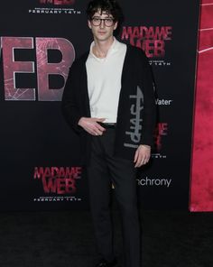 a man wearing glasses and a white shirt standing in front of a red carpeted wall