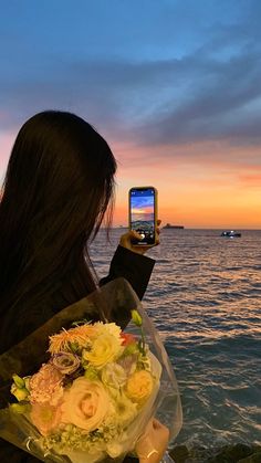 a woman taking a photo with her cell phone on the beach while holding a bouquet of flowers