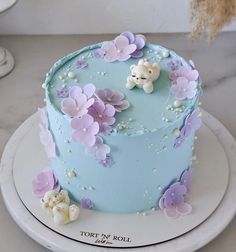 there is a blue cake with purple flowers on the side and a teddy bear on top